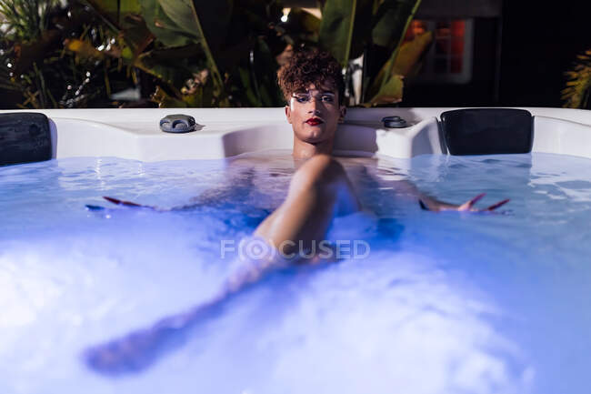 Giovane uomo transessuale con lunghe unghie guardando la fotocamera sdraiato in vasca idromassaggio al crepuscolo — Foto stock