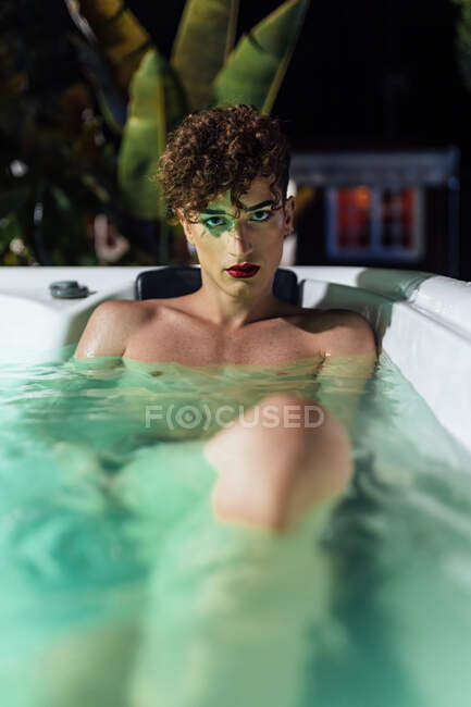 Jovem transexual com unhas longas olhando para a câmera deitada na banheira de hidromassagem ao entardecer — Fotografia de Stock