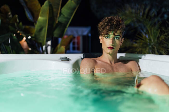 Junger transsexueller Mann mit langen Nägeln blickt in der Abenddämmerung in die Kamera, die im Whirlpool liegt — Stockfoto