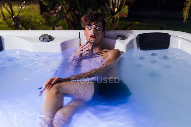 Giovane uomo transessuale con le unghie lunghe e gli occhi chiusi sdraiati in vasca idromassaggio al tramonto — Foto stock