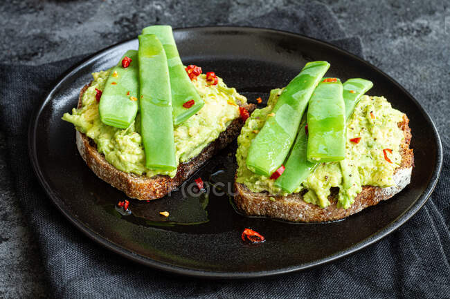 Pane tostato appetitoso con guacamole fresco e baccelli di piselli verdi guarniti con peperoni rossi e serviti su piatto nero — Foto stock