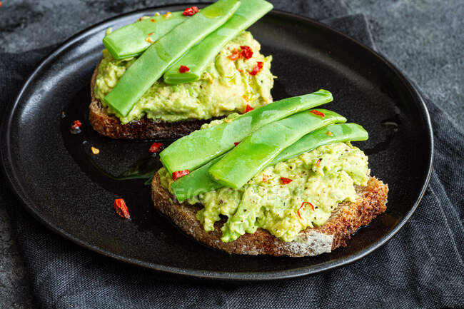 Appetizing toasts with fresh guacamole and green peas pods garnished with red peppers and served on black plate — Stock Photo