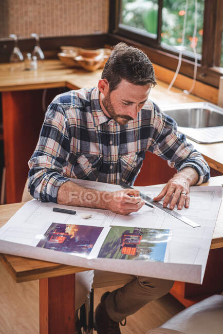 Серьезный мужчина-архитектор, сидящий за столом на кухне и рисующий чертеж здания карандашом и линейкой, работая дома — стоковое фото
