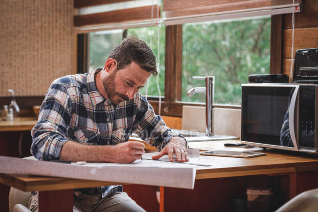 Seriöser männlicher Architekt sitzt in der Küche am Tisch und zeichnet mit Bleistift und Lineal Baupläne für das Haus, während er zu Hause arbeitet — Stockfoto