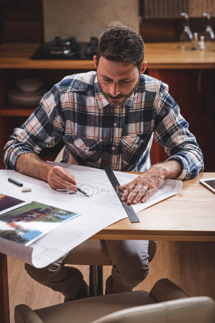 Arquitecto masculino serio sentado a la mesa en la cocina y dibujando el plano del edificio con lápiz y regla mientras trabaja en casa - foto de stock