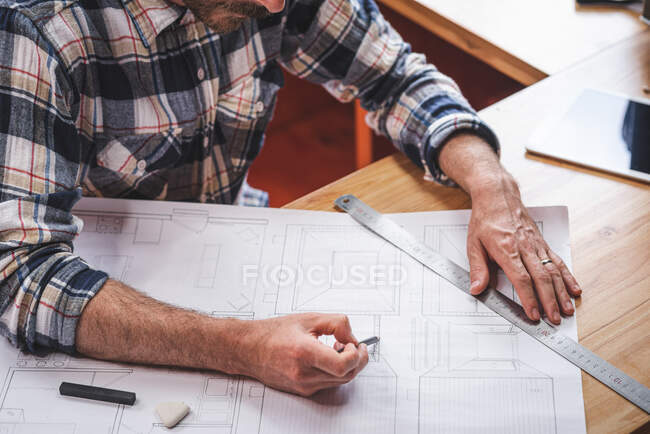 Anonymer männlicher Architekt sitzt am Tisch in der Küche und zeichnet mit Bleistift und Lineal den Bauplan für das Gebäude, während er zu Hause arbeitet — Stockfoto