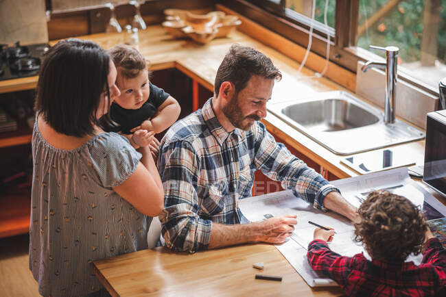 Hoher Winkel des lächelnden männlichen Architekten am Tisch sitzend und Entwurf des Hauses in der Küche mit Familie — Stockfoto