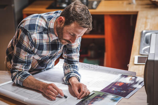Encantado arquitecto masculino de pie a la mesa y plano de dibujo de la casa en papel mientras trabaja en el proyecto en casa - foto de stock