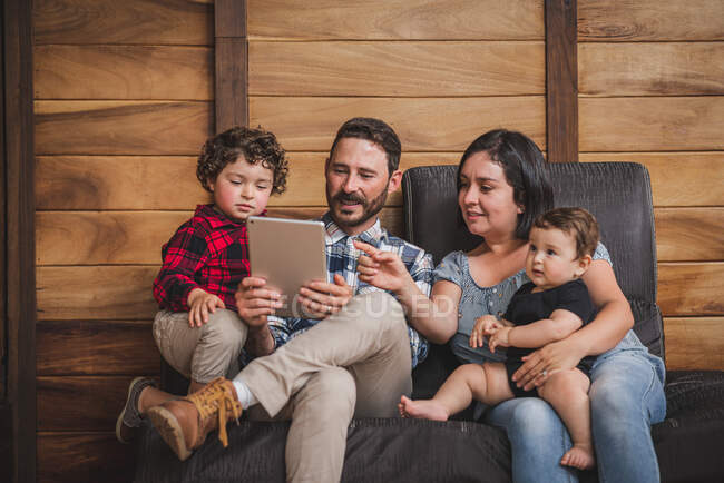 Happy mother and father sitting on sofa with kids and watching video together on tablet at home — Stock Photo