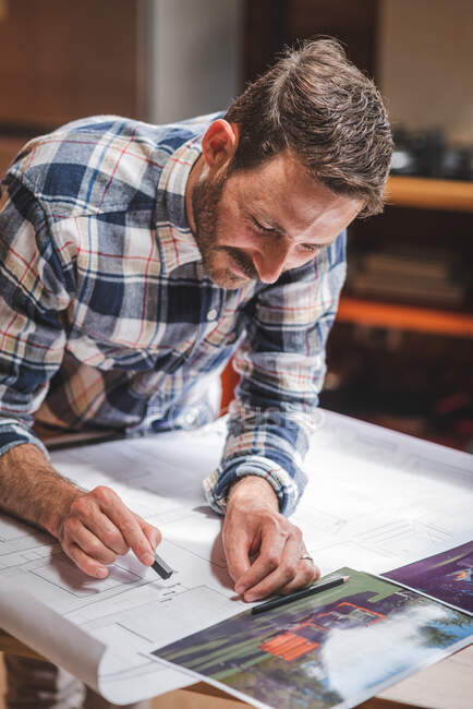 Entzückter männlicher Architekt steht am Tisch und zeichnet Plan des Hauses auf Papier, während er zu Hause an einem Projekt arbeitet — Stockfoto