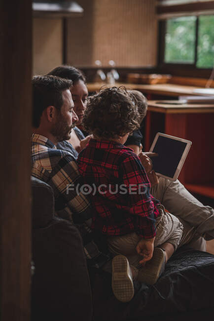 Mãe e pai felizes sentados no sofá com crianças e assistindo vídeo juntos no tablet em casa — Fotografia de Stock