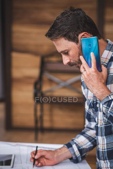 Side view of focused male architect drawing blueprint of house and speaking on smartphone while standing at table and working from home — Stock Photo