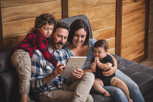 Happy mother and father sitting on sofa with kids and watching video together on tablet at home — Stock Photo