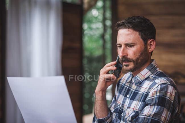 Architecte masculin concentré avec un plan parlant sur un téléphone portable tout en travaillant à la maison et en discutant d'un nouveau projet — Photo de stock