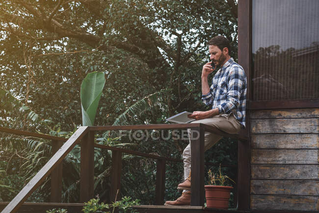 Side view of serious male architect sitting on railing on wooden terrace and talking on smartphone while discussing project — Stock Photo