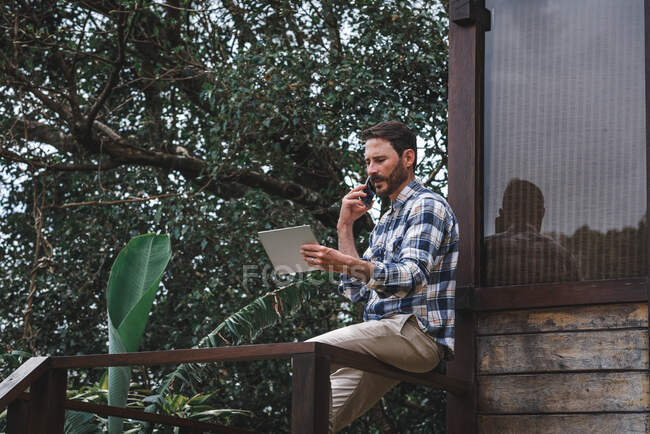 Vista lateral do arquiteto masculino sério sentado no corrimão no terraço de madeira e falando no smartphone enquanto discute o projeto — Fotografia de Stock