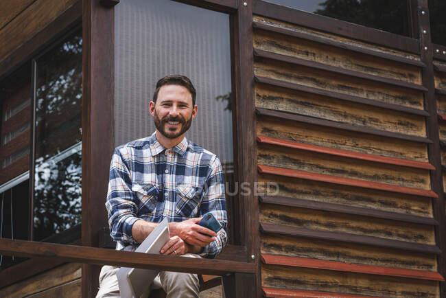 Bajo ángulo de sonriente arquitecto masculino con borradores de papel y dispositivos sentados en barandilla en la terraza de la casa de madera moderna y mirando a la cámara - foto de stock