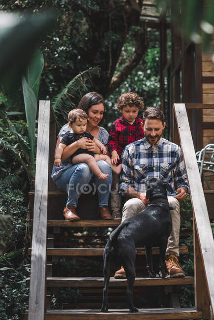 Fröhliches Paar mit Kindern sitzt mit Hund auf Holztreppe des modernen Hauses — Stockfoto