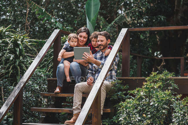 Cheerful couple with cute children sitting on wooden steps of house and taking selfie on tablet — Stock Photo