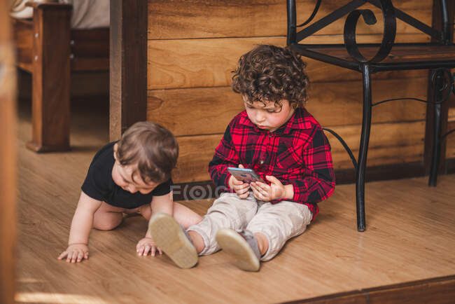 Чарівний хлопчик переглядає смартфон, сидячи на підлозі біля маленького брата вдома — стокове фото