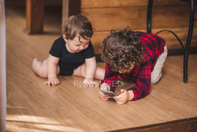 Adorabile smartphone di navigazione ragazzo mentre seduto sul pavimento vicino a fratellino a casa — Foto stock