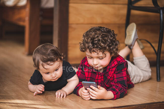 Очаровательный мальчик просматривает смартфон, лежа на полу рядом с маленьким братом и сестрой дома — стоковое фото