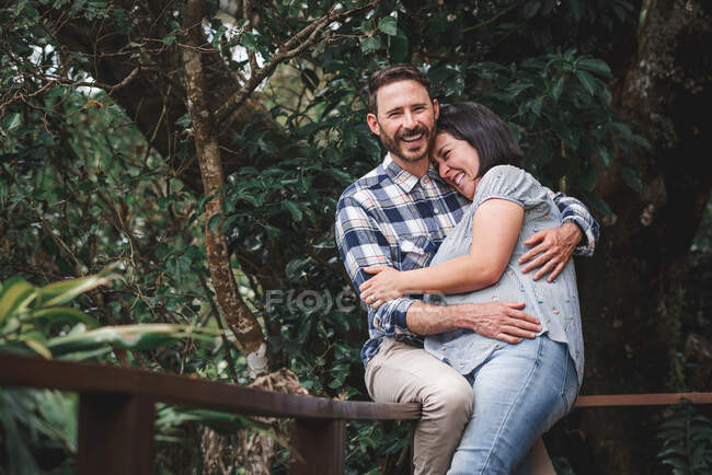 Cheerful couple in love embracing on wooden terrace of house in woods and laughing — Stock Photo