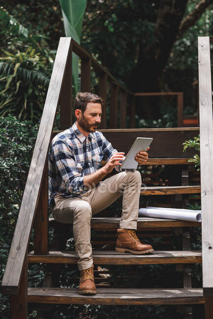 Arquiteto masculino focado usando tablet enquanto sentado em degraus de madeira da casa e trabalhando em projeto remoto — Fotografia de Stock