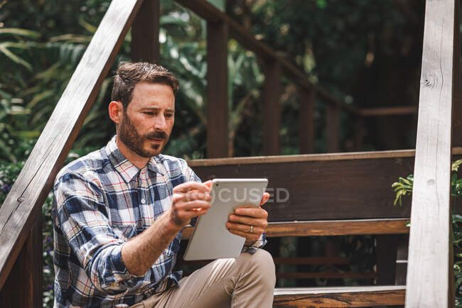 Konzentrierter männlicher Architekt mit Tablet, während er auf hölzernen Stufen des Hauses sitzt und an einem entfernten Projekt arbeitet — Stockfoto