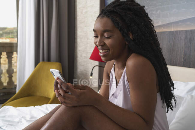 Счастливая молодая афроамериканка с длинными кудрявыми волосами во сне улыбается и читает сообщение на смартфоне, отдыхая на удобной кровати — стоковое фото