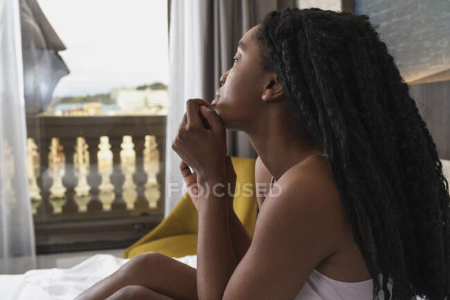Бічний вид приваблювання молодих босоногих чорних жінок тисячоліття з африканськими плечима в стильний сонцезахисний одяг сидячи на м'якому ліжку і озираючись геть — стокове фото
