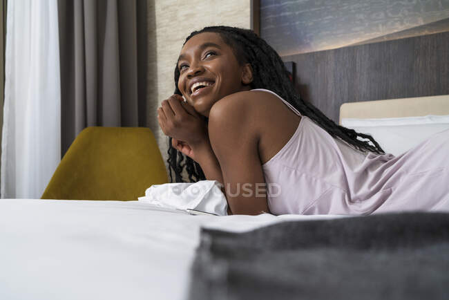 Atractivo joven descalzo hembra negra millennial con trenzas afro en ropa de dormir con estilo acostado en la cama suave y mirando hacia otro lado - foto de stock