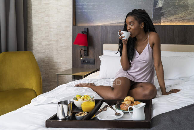Corpo pieno di contenuti giovane afroamericana turista donna in camicia da notte seduta su un comodo letto con vassoio di deliziosa colazione e bere caffè in hotel moderno guardando altrove — Foto stock