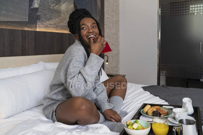 Corpo inteiro de conteúdo jovem afro-americano turista em roupa de noite sentado em cama confortável com bandeja de delicioso café da manhã no hotel moderno olhando para longe — Fotografia de Stock