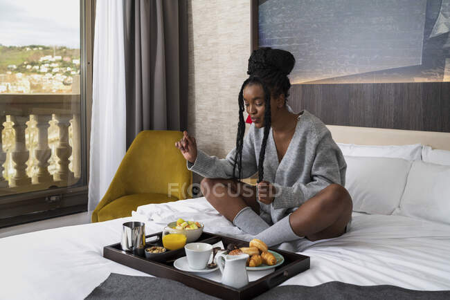 Corpo pieno di contenuti giovane afroamericana turista donna in camicia da notte seduta su un comodo letto con vassoio di deliziosa colazione in hotel moderno — Foto stock