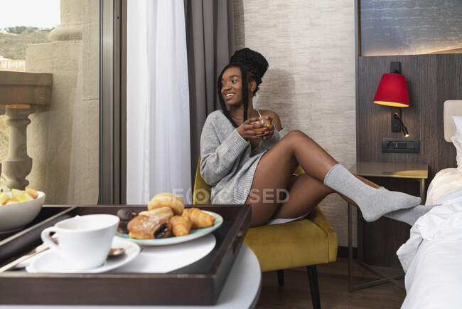 Longitud completa de viajero femenino étnico joven positivo sentado en la silla en la habitación del hotel y beber jugo fresco durante el desayuno en el día soleado - foto de stock