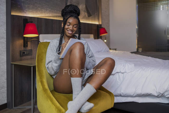 Verträumte junge positive Afroamerikanerin in lässiger Kleidung und Socken, die nachdenklich auf einem bequemen Sessel sitzt, während sie im gemütlichen Schlafzimmer Notizen macht — Stockfoto