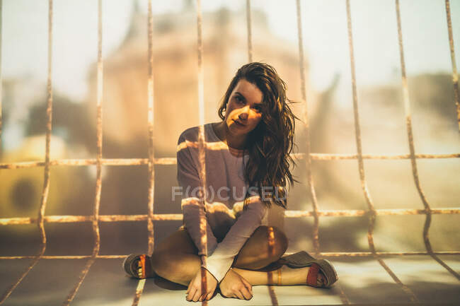 Jovem fêmea em roupa de verão olhando para a câmera enquanto sentada de pernas cruzadas sob projeção de barras de gaiola — Fotografia de Stock