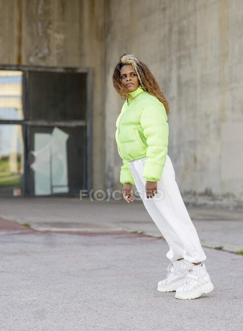 Ganzkörperansicht einer jungen Afroamerikanerin mit gefärbten lockigen Haaren, die eine elegante grüne Jacke mit weißen Hosen und trendigen Stiefeln trägt, während sie auf einer gepflasterten städtischen Straße steht und wegschaut — Stockfoto