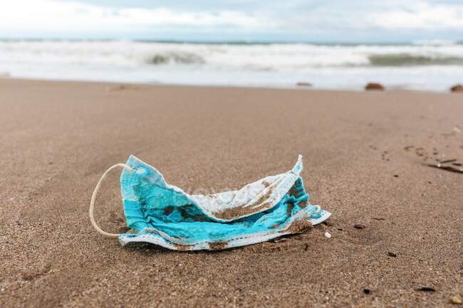 Gebrauchte Schutzmaske am Strand, angespült von Meereswellen, die das Konzept der Umweltverschmutzung mit medizinischen Abfällen aufzeigt — Stockfoto