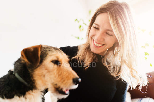 Mulher loura alegre acariciando focinho de cão de raça pura amigável enquanto descansa na cama em casa — Fotografia de Stock