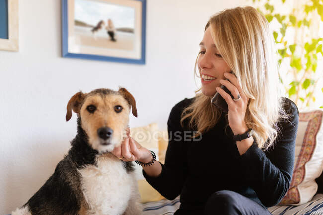 Aufrichtige blonde Hündin streichelt entzückenden reinrassigen Hund, während sie auf dem Bett im Haus mit dem Handy spricht — Stockfoto
