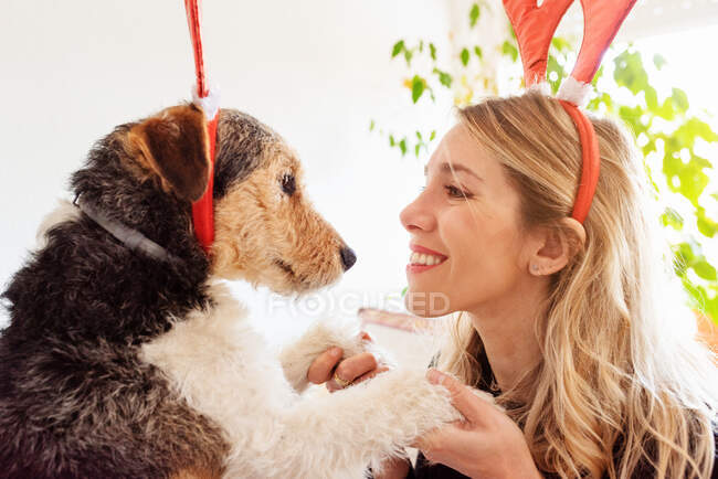 Side view of happy female in decorative deer horns cuddling adorable purebred dog on bed — Stock Photo