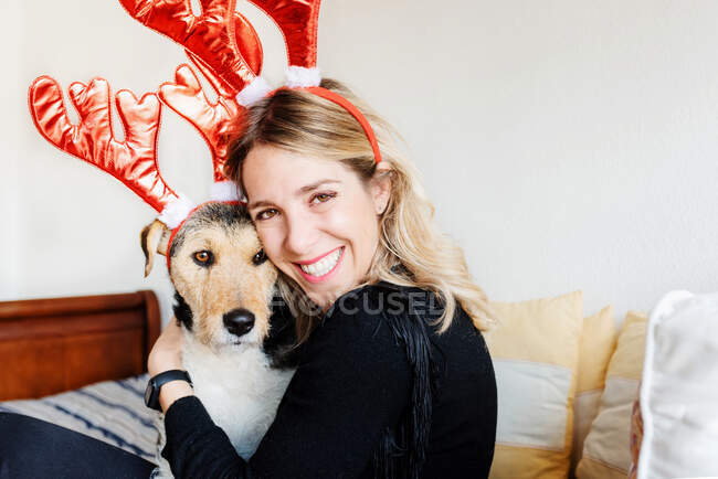 Happy female in decorative deer horns cuddling adorable purebred dog while looking at camera on bed — Stock Photo