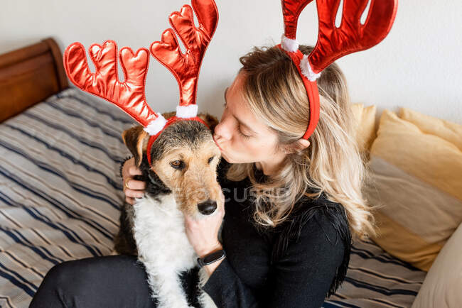 Mulher feliz em chifres de veado decorativos abraçando adorável cão de raça pura na cama — Fotografia de Stock