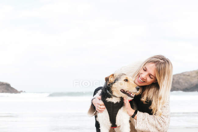 Vista lateral de la mujer abrazando lindo perro de raza pura mientras se miran el uno al otro contra el mar bajo el cielo nublado - foto de stock