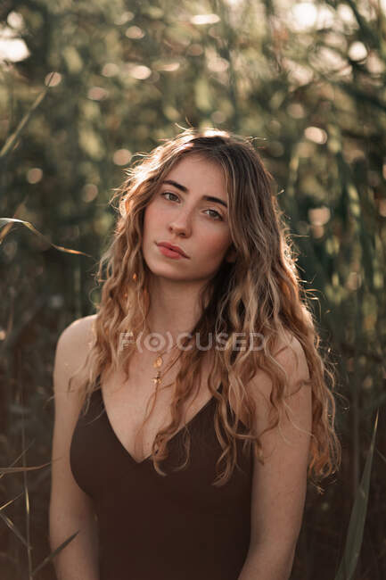 Charming young brunette with long wavy hair wearing black swimsuit looking at camera while standing against blurred green trees in sunny garden — Stock Photo