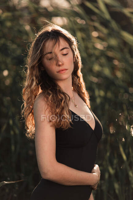 Vista lateral de la encantadora morena joven con el pelo ondulado largo con traje de baño negro con los ojos cerrados de pie contra los árboles verdes borrosos en el jardín soleado - foto de stock