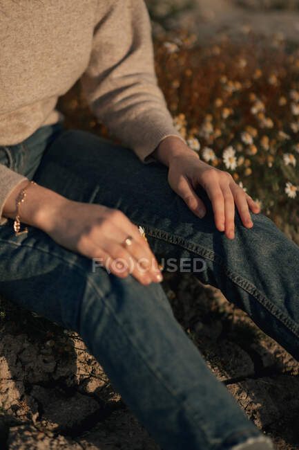 Recortado viajero femenino irreconocible en ropa casual sentado solo en el prado floreciente contra el campo arado borroso mientras se relaja en el campo en el día de primavera - foto de stock