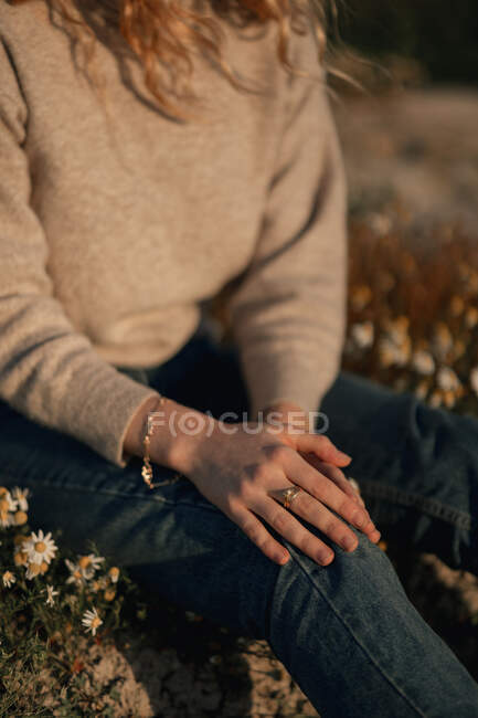 Cropped unrecognizable female traveler in casual wear sitting alone on flowering meadow against blurred plowed field while relaxing in countryside in spring day — Stock Photo